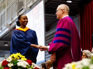 A woman wearing a blue convocation gown and a yellow hood shakes the hand of a man wearing a magenta gown.