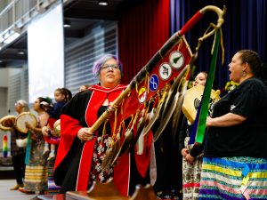 A woman wearing a ceremonial gown carries an Indigenous staff across a stage at Brock University.