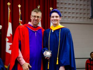 A man wearing a red ceremonial gown presents a medal to another man wearing a blue ceremonial gown during Brock University’s 115th Convocation ceremony.