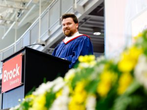 A man wearing a blue convocation gown speaks at a podium with a red Brock University logo on stage inside Ian Beddis Gymnasium during a graduation ceremony.