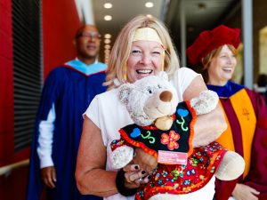 A smiling woman carries a ceremonial teddy bear through the halls of Brock University’s Walker Complex before a graduation ceremony. She is followed by people wearing academic robes.