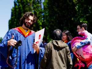 A man wearing a blue gown smiles and gives a thumbs up while holding a degree outdoors on a sunny day following a university graduation ceremony.