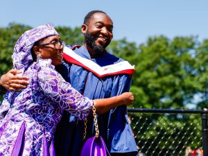 A man wearing a blue robe and white hood smiles while embracing a woman wearing purple outside with a backdrop of green trees and blue skies after a university graduation ceremony.