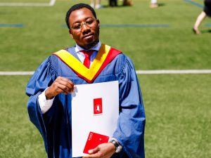 A man wearing a blue gown and yellow hood smiles while holding a degree from Brock University while outside on an athletic field.