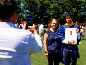 A man in a convocation gown and a woman wearing purple pose for a photograph being taken by a man holding a mobile phone outside after a university graduation ceremony.