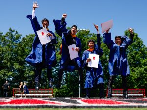 A group of four men wearing blue convocation gowns jump into the air in celebration outside on a turf field at Brock University after a graduation ceremony.