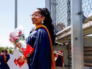 A woman wearing glasses and a blue graduation gown holds red flowers and a degree from Brock University.