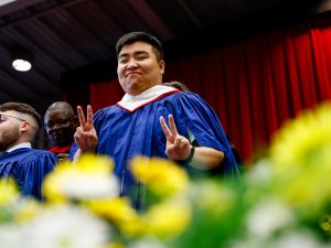 A man wearing a ceremonial gown holds up the peace sign with his fingers standing behind a row of flowers.