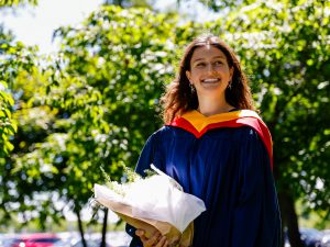 A woman wearing a blue gown and a yellow hood holds flowers while smiling, standing outdoors amongst the sun-lit trees on Brock University’s campus.