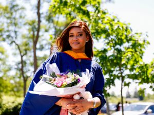 A woman wearing a blue gown and a yellow hood holds flowers while smiling, standing outdoors the sun-lit trees on Brock University’s campus.