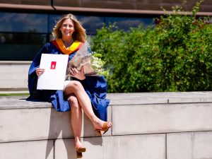 A smiling woman wearing a ceremonial blue gown sits on a concrete ledge in front of Brock University while holding a degree and flowers.