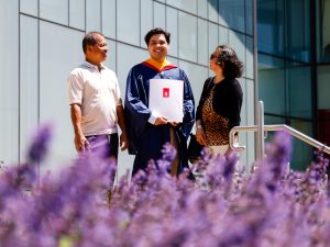 A man in a blue academic robe and holding a diploma stands between two loved ones outside a university graduation ceremony.