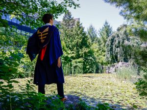 A man wearing a ceremonial blue convocation gown stands on the edge of a pond surrounded by shade and green trees in the heart of Brock University’s main campus.