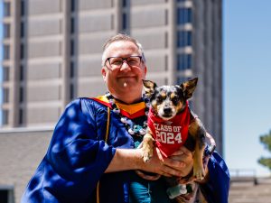 A man wearing a glasses and ceremonial blue robe smiles outdoors while holding a little dog wearing a red Class of 2024 scarf with Brock University’s Schmon Tower in the background.