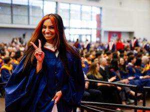 A woman wearing a ceremonial gown walks in front of a sea of graduates with a bright smile and holding up the peace sign with her fingers.