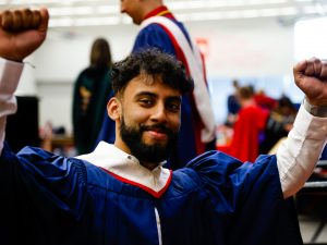 A man wearing a ceremonial gown holds up his arms in celebration in Brock University’s gym during a graduation ceremony.