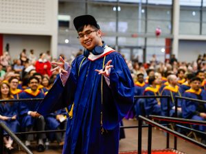 A man wearing a ceremonial gown gestures in a celebratory manner with arms wide open in front of a sea of graduates at Brock University’s 115th Convocation Ceremony.