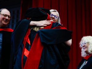 Two women in academic robes hug on stage at Brock University’s Ian Beddis Gymnasium.