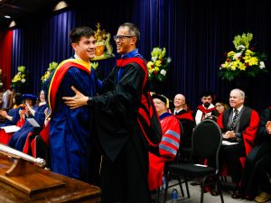 A father embraces his son at the end of the graduation stage at Brock University. Both are wearing academic robes.