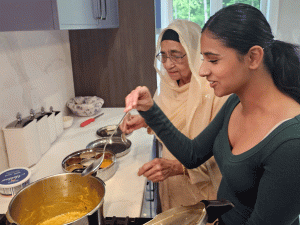 Honours Bachelor of Public Health graduate Gehreen Brar holds a metal spoon that she is dipping into a pot on the stove while her grandmother Lakhwinder Kaur Brar looks on.