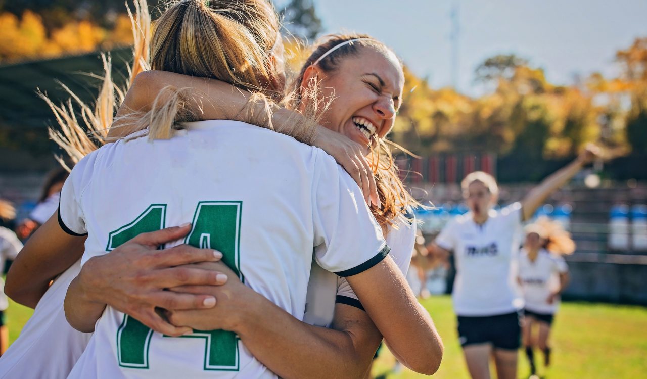 A group of female soccer players hug and celebrate on a soccer pitch.