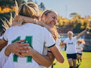 A group of female soccer players hug and celebrate on a soccer pitch.