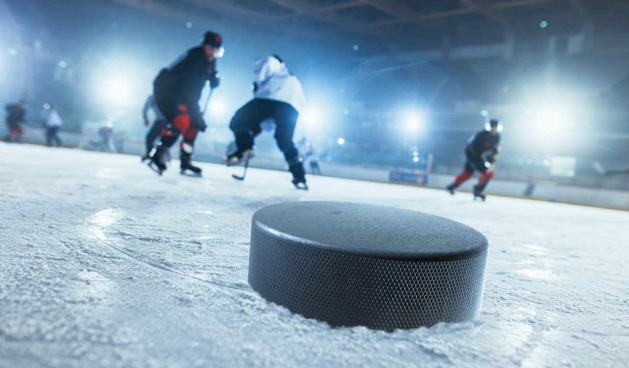 Close-up photo of hockey puck on ice hockey rink and hockey players trying to get the puck in the background.