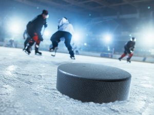 Close-up photo of hockey puck on ice hockey rink and hockey players trying to get the puck in the background.