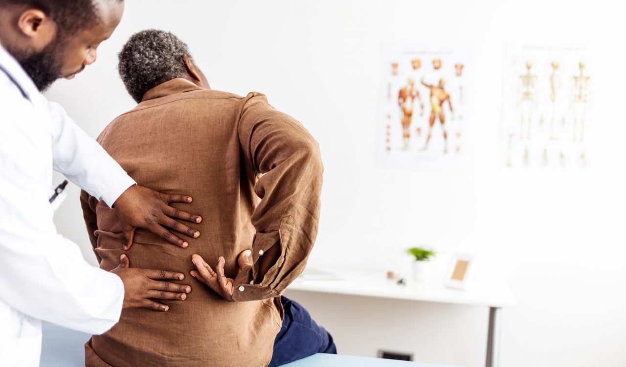 A doctor examines the lower back of a patient who is sitting on a bed in a doctor's office.