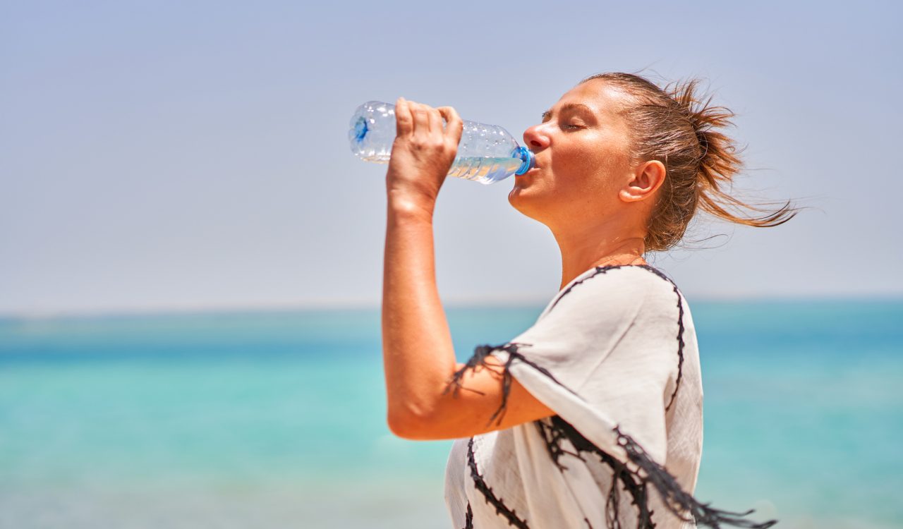 A woman drinks from a water bottle while standing beside the ocean on a hot day.