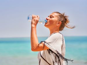 A woman drinks from a water bottle while standing beside the ocean on a hot day.