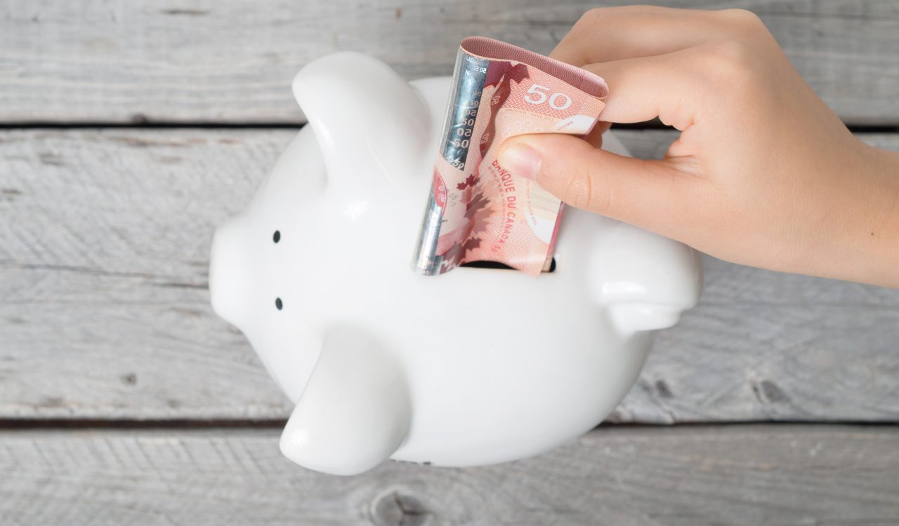 A hand inserts a $50 bill into a white piggy bank against a wooden grey background.