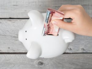 A hand inserts a $50 bill into a white piggy bank against a wooden grey background.