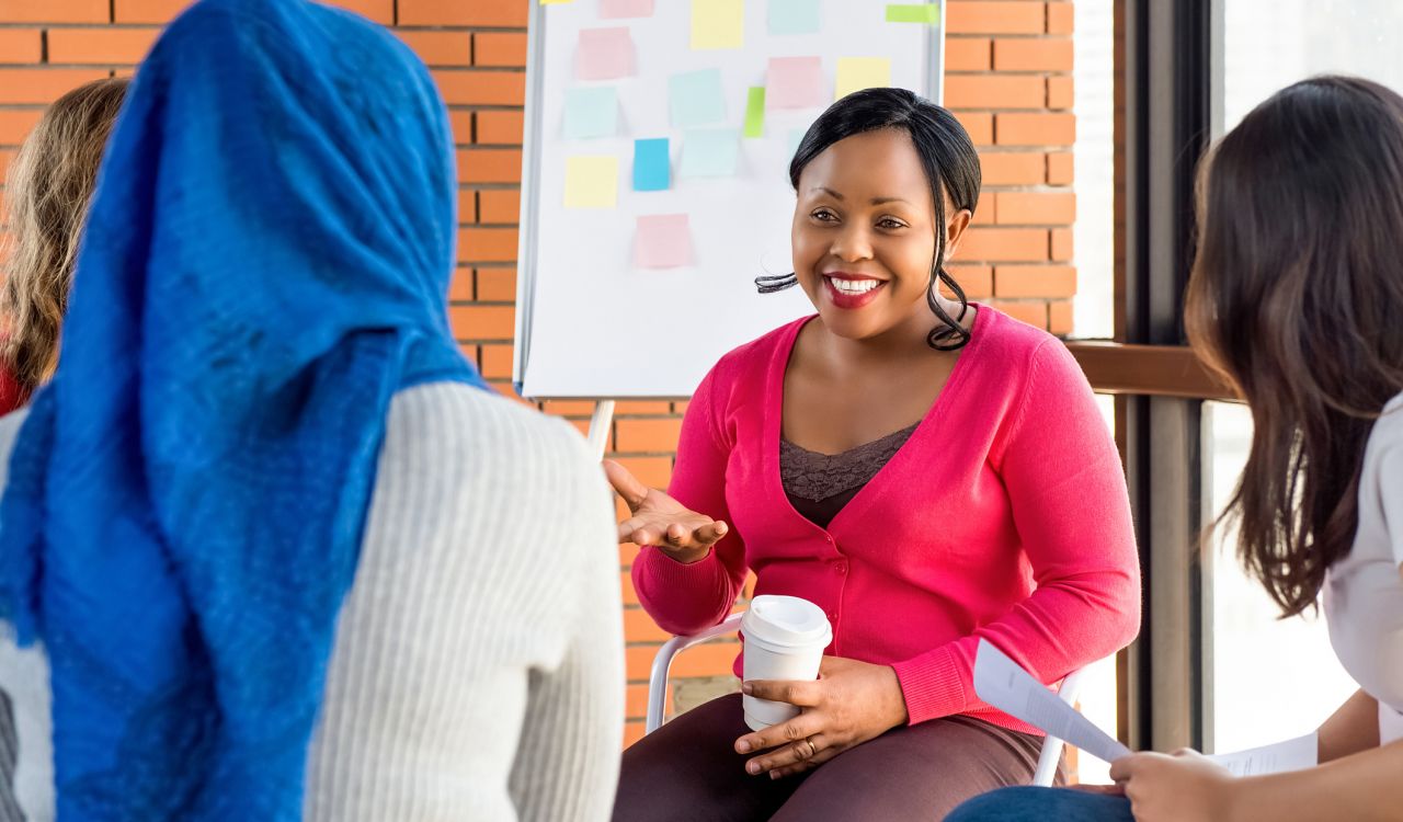 A group of four diverse women sitting in chairs in a circle. In the foreground is the back of a woman wearing a blue head covering. Facing the camera is a smiling woman holding a cup of coffee and gesturing with her hand, with side views of the other two women.