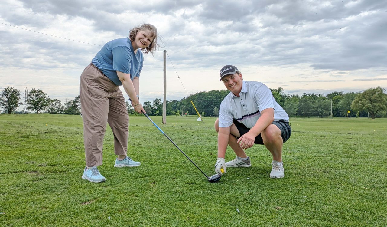 A person stands ready to swing a golf club on a golf green while a second is crouched down holding a golf ball and providing direction.