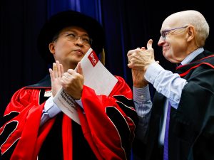 Two men in academic regalia applaud on stage during a university graduation ceremony.