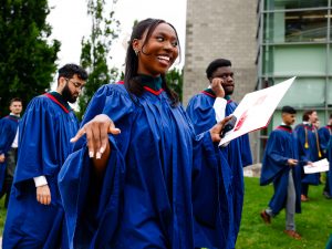 A woman in a blue academic robe holds a diploma and smiles after a university graduation ceremony.