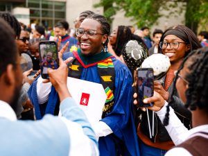 A new university graduate in a blue academic robe holds a diploma and poses for a photo taken by a loved one.