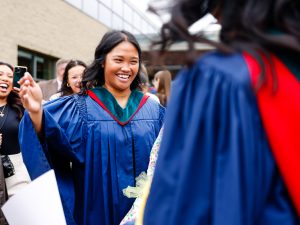 Two young women in blue academic robes hug outside after a university graduation ceremony.