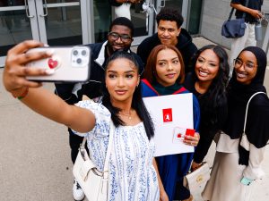 A young woman in a blue academic robe poses for a selfie with loved ones while holding her diploma after a university graduation ceremony.