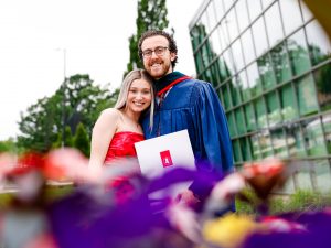 A man in a blue academic robe poses for a photo with a loved one while holding his diploma after a university graduation ceremony.