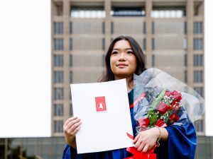 A young woman in a blue academic robe poses with flowers and a diploma after a university graduation ceremony.