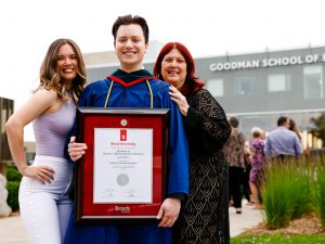 A young man in a blue academic robe holds a framed diploma and poses for a photo with two loved ones after a university graduation ceremony.