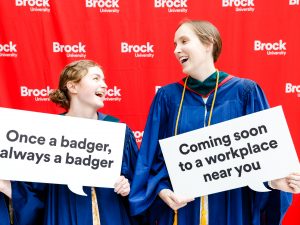 Two young women in blue academic robes pose for photo while holding speech bubble props after a university graduation ceremony.