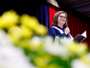 A woman in blue academic robes speaks into a microphone on stage during a university graduation ceremony.