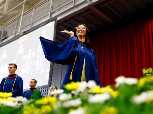 A woman in a blue academic robe waves while crossing the stage during a university graduation ceremony.