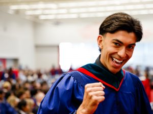 A man in a blue academic robe holds up a fist and smiles while crossing the stage during a university graduation ceremony.