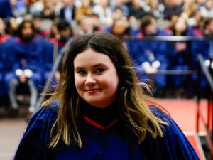 A woman in a blue academic robe smiles while leaving the stage during a university graduation ceremony.