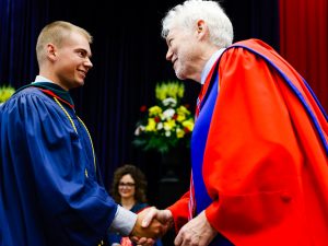 A man in a blue academic robe shakes hands with a man in red and blue academic regalia during a university graduation ceremony.