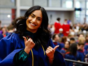 A woman in a blue academic robe smiles and gives two thumbs up while leaving the stage during a university graduation ceremony.